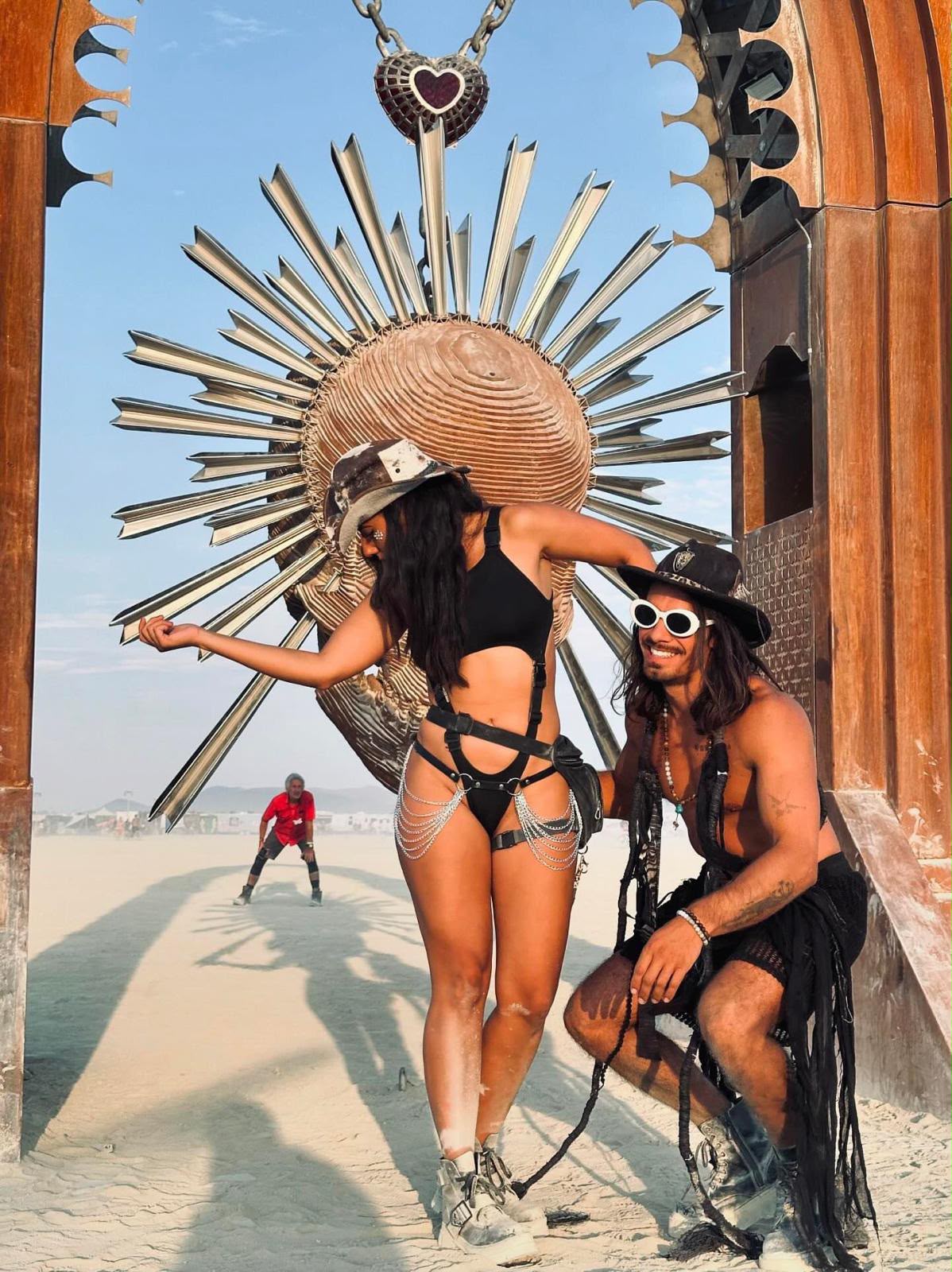 Vista Collective burners under a metal sun arch at Burning Man in leather harness and chain skirt